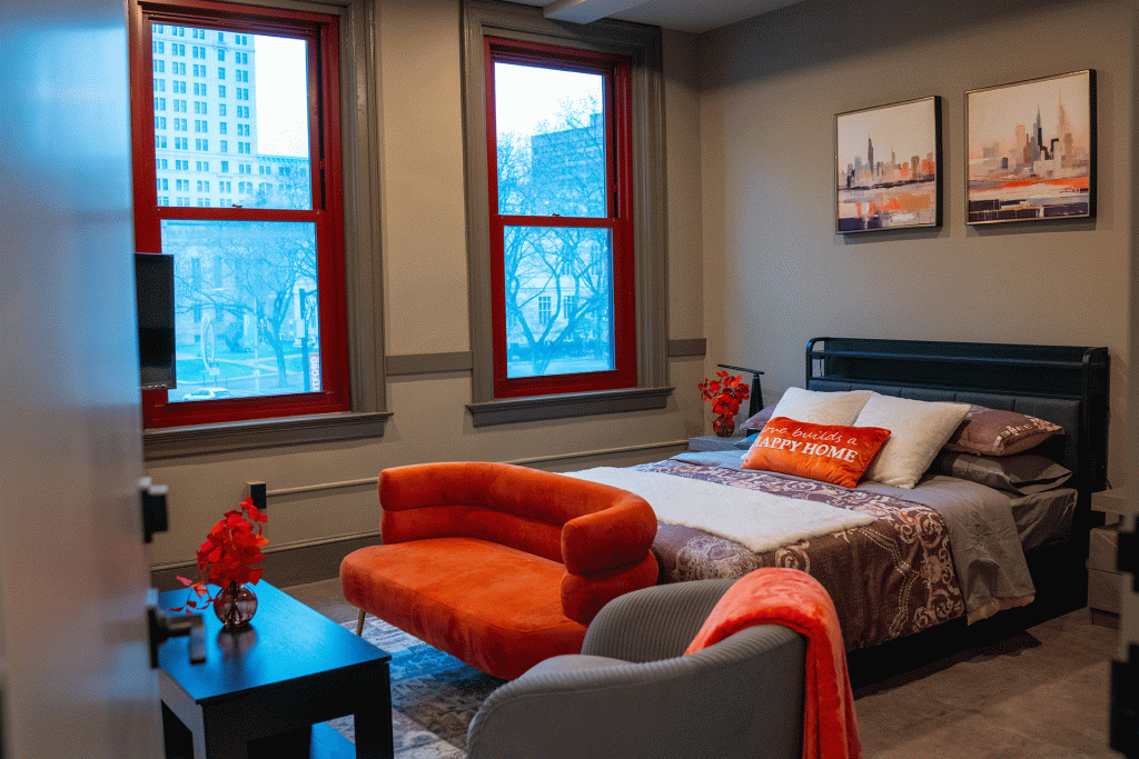Bedroom with red-framed windows, city skyline view, orange velvet bench, and gray armchair beside the bed