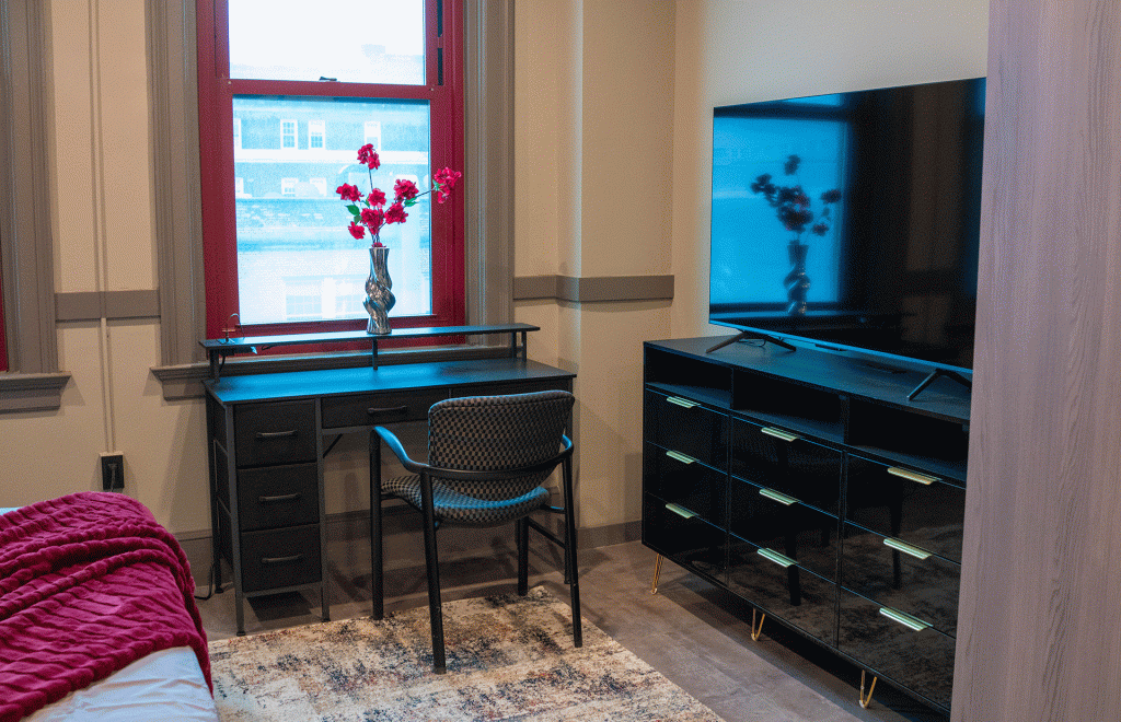 Bedroom desk with chair and flower vase beside a red-framed window, plus a mounted TV on a dresser